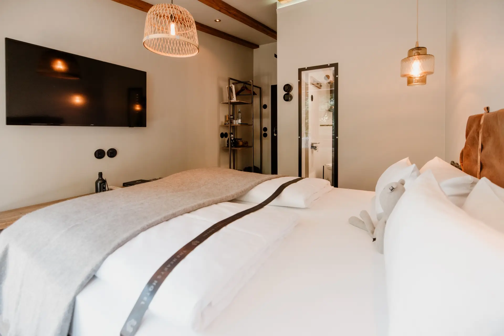 Gemütliches Schlafzimmer mit Doppelbett, TV, hölzernem Regal und Blick auf das Badezimmer durch Glastür. Warmes Licht, entspannte Atmosphäre.