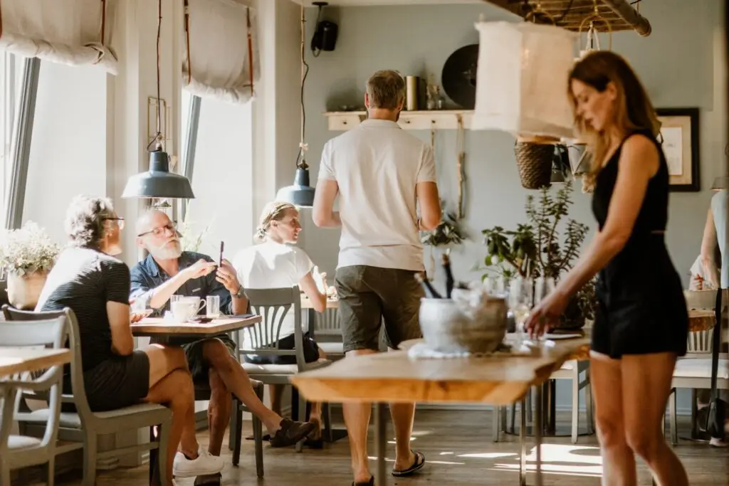 Freundliche Atmosphäre in einem Café, Gäste sitzen und unterhalten sich, während eine Servicekraft am Tisch vorbeigeht.