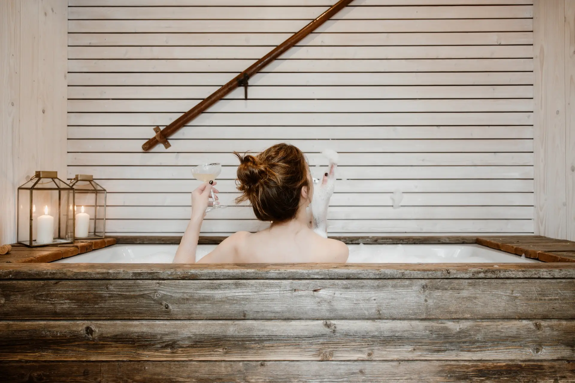 Frau sitzt in einer Holzbadewanne, hält ein Glas und zeigt mit der anderen Hand Seifenblasen. Kerzen und eine Holzverkleidung im Hintergrund.