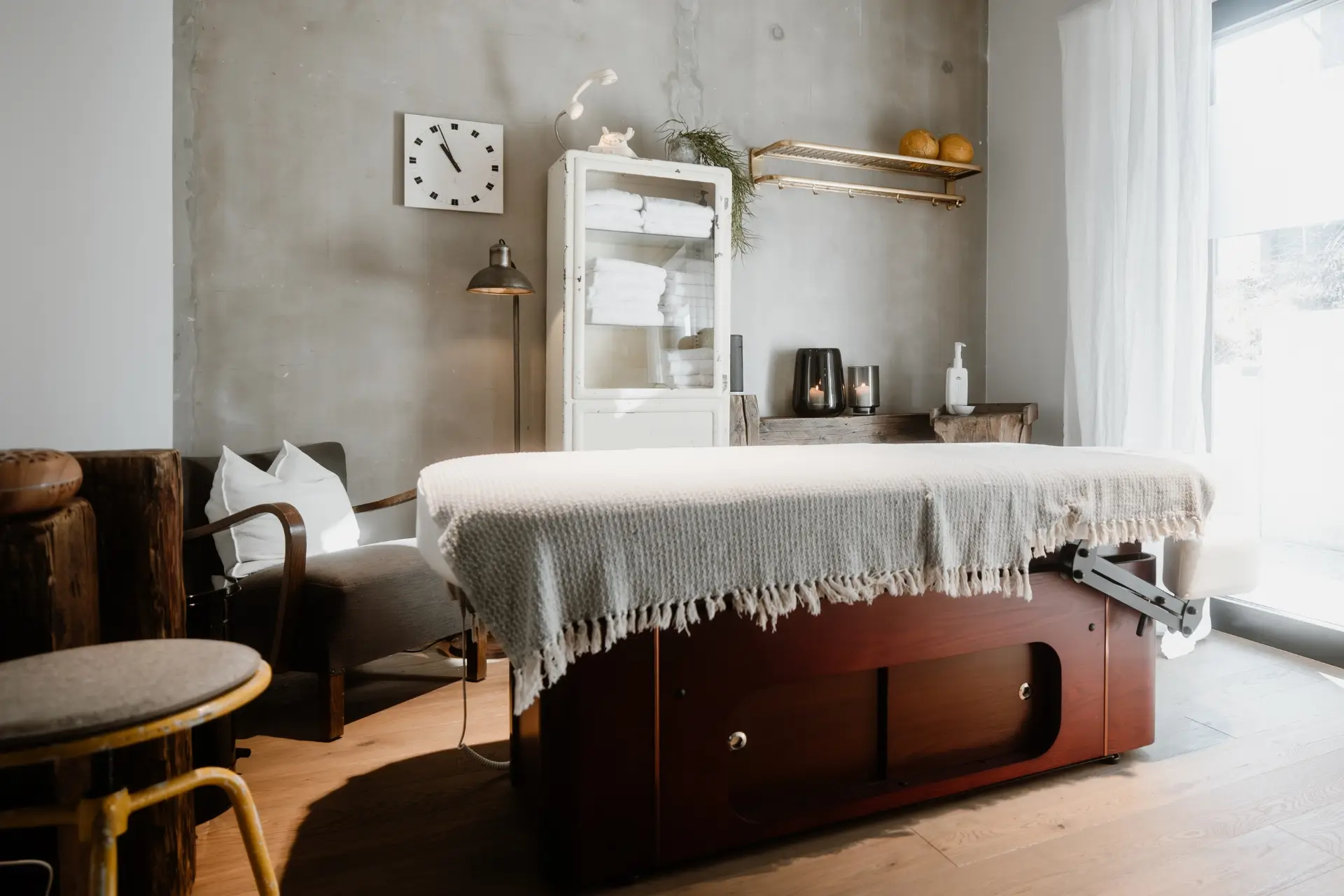 Wellnessraum mit Massagetisch, Sitzgelegenheiten, handtuchgefülltem Regal und sanfter Beleuchtung. Helle, einladende Atmosphäre.