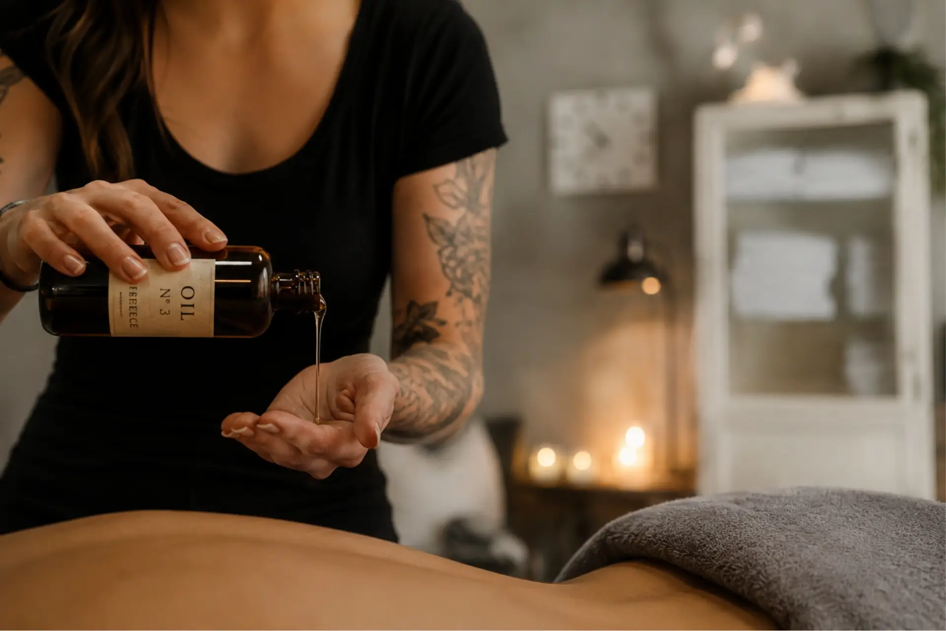 Masseurin gießt Massageöl in die Hand, während eine Person auf einer Liege liegt. Der Raum ist stilvoll und entspannend eingerichtet.