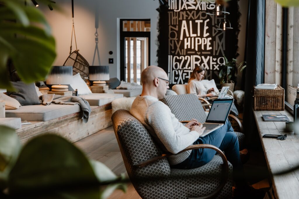 Mann mit Brille arbeitet am Laptop, während eine Frau im Hintergrund ebenfalls tippt. Stilvolles, modernes Café-Ambiente.