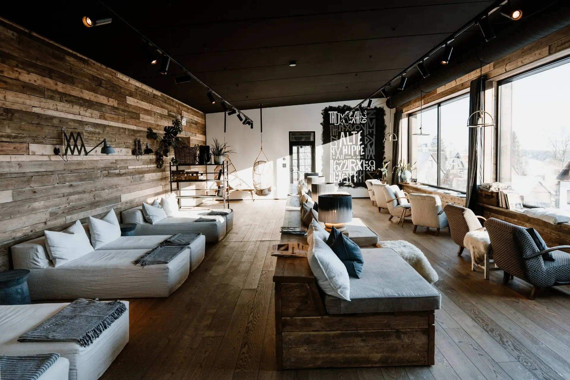 Modernes, helles Lounge-Ambiente mit Sitzgelegenheiten, Holzverkleidung und großen Fenstern, die Tageslicht hereinkommen lassen.
