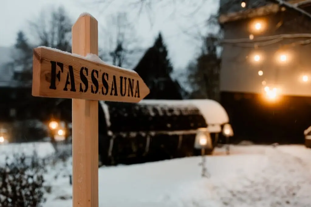 Holzschild weist auf die Fasssauna in einer schneebedeckten Umgebung, mit sanfter Beleuchtung im Hintergrund.