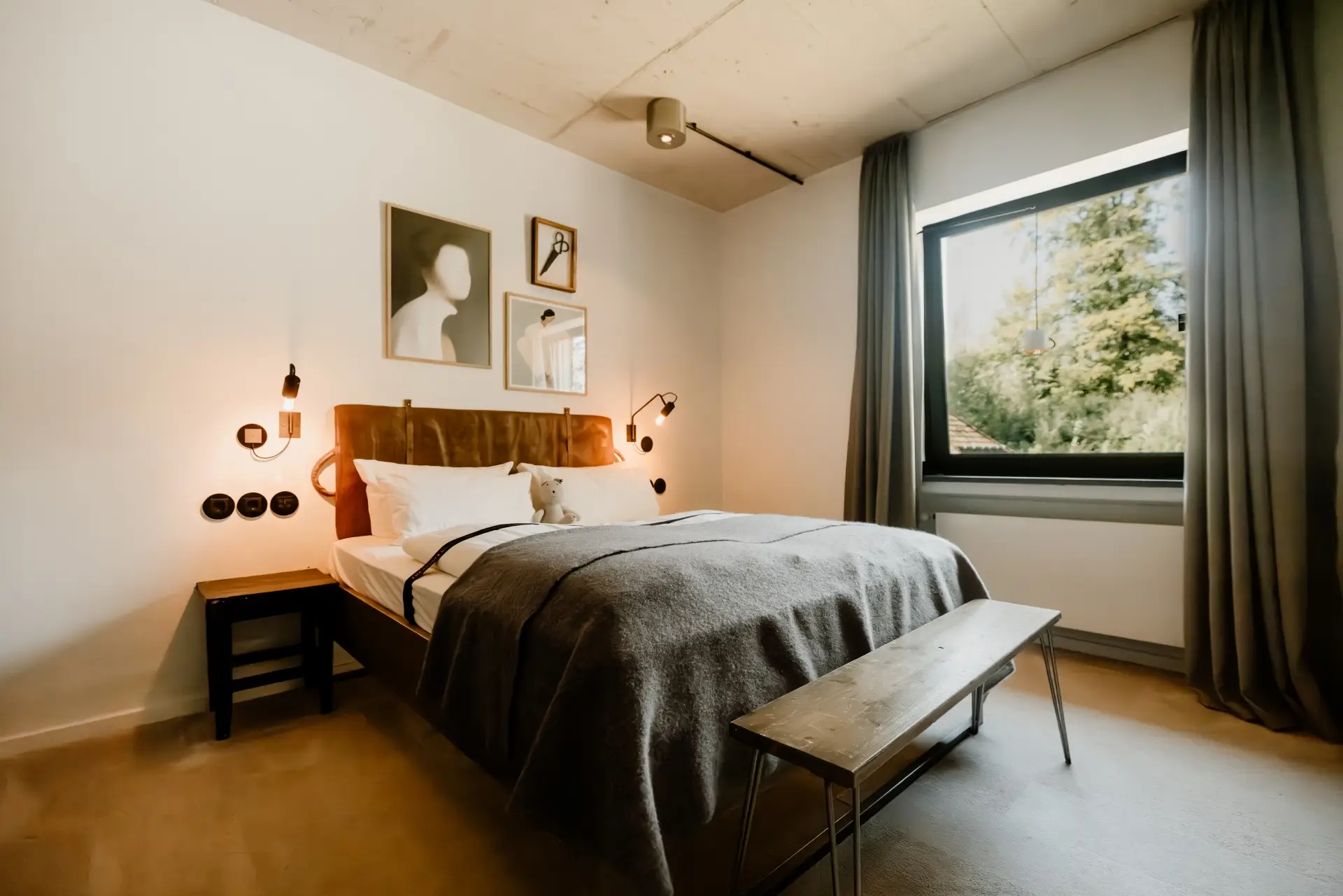 Moderne Schlafzimmer mit einem Doppelbett, grauer Decke, Tisch und Fenster mit Blick auf Bäume. Kunstwerke an der Wand.