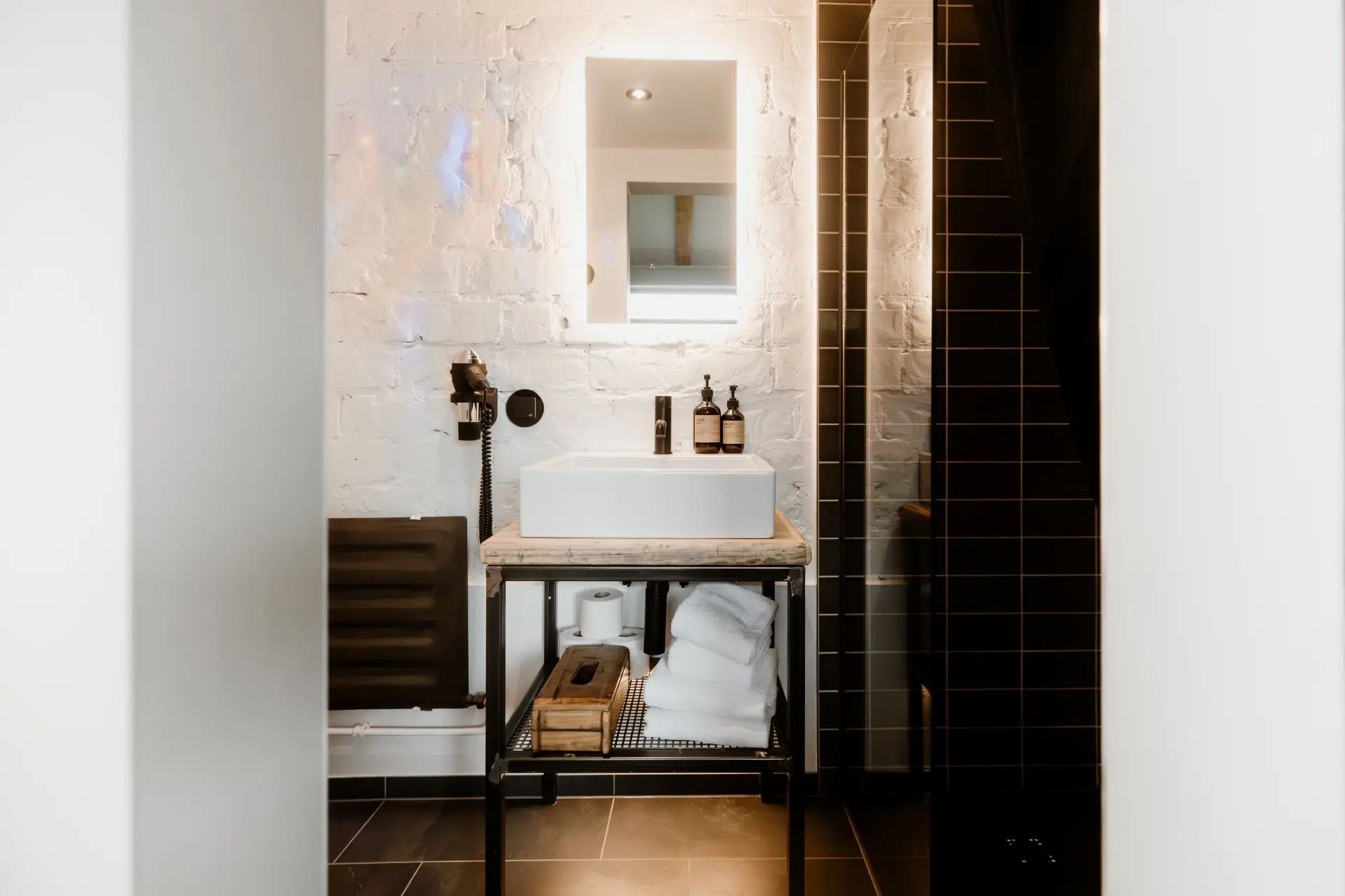 Badezimmer mit einem Waschbecken auf einem Holzgestell, Handtüchern und Produkten, beleuchtet von einem Wandspiegel.