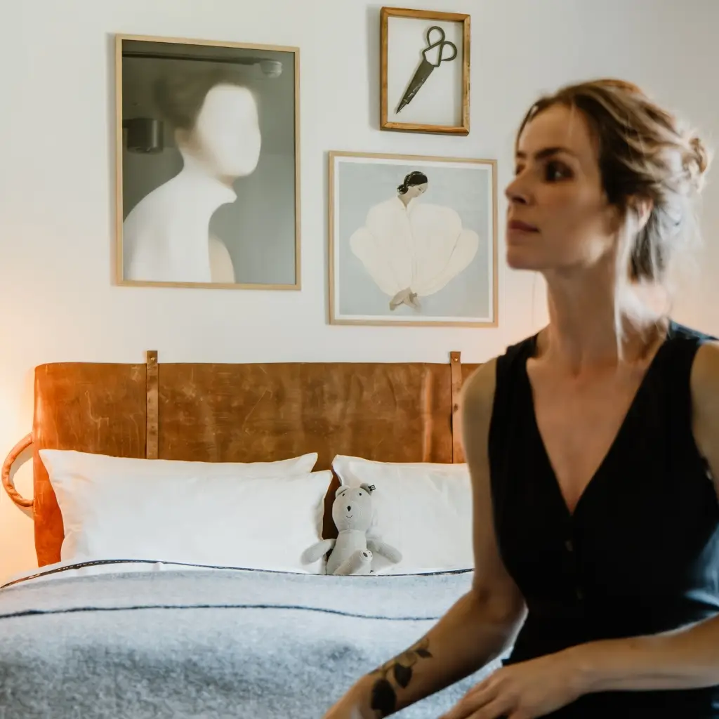 Frau sitzt auf dem Bett in einem hellen Zimmer, umgeben von Kunstwerken, mit einem Kuscheltier im Vordergrund.