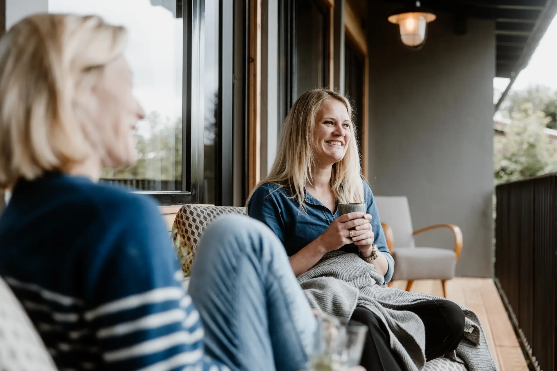 Zwei Frauen sitzen entspannt auf einer Veranda, die eine hält eine Tasse in der Hand, die andere schaut lächelnd.