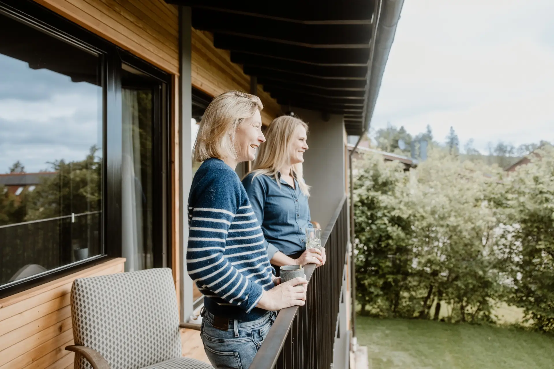 Zwei Frauen stehen auf einem Balkon, halten Getränke in der Hand und blicken entspannt in die Landschaft.