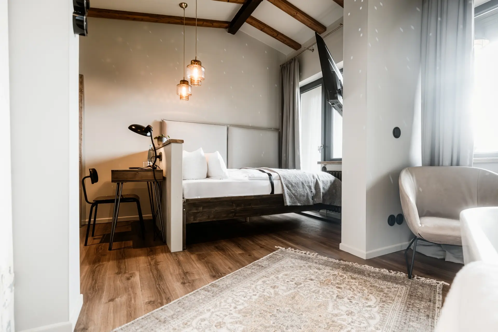 Modernes Zimmer mit Doppelbett, Arbeitsplatz und Sitzbereich. Große Fenster mit Vorhängen, Holzdecken und stilvollem Teppich.