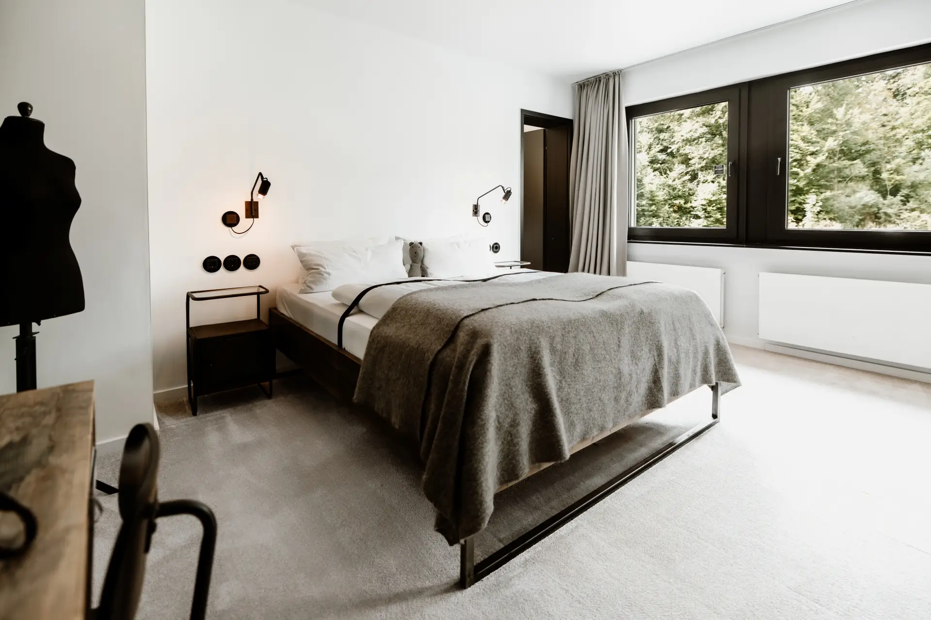 Modernes Schlafzimmer mit einem Doppelbett, Nachttisch, Leseleuchte und Fenster mit Blick ins Grüne.