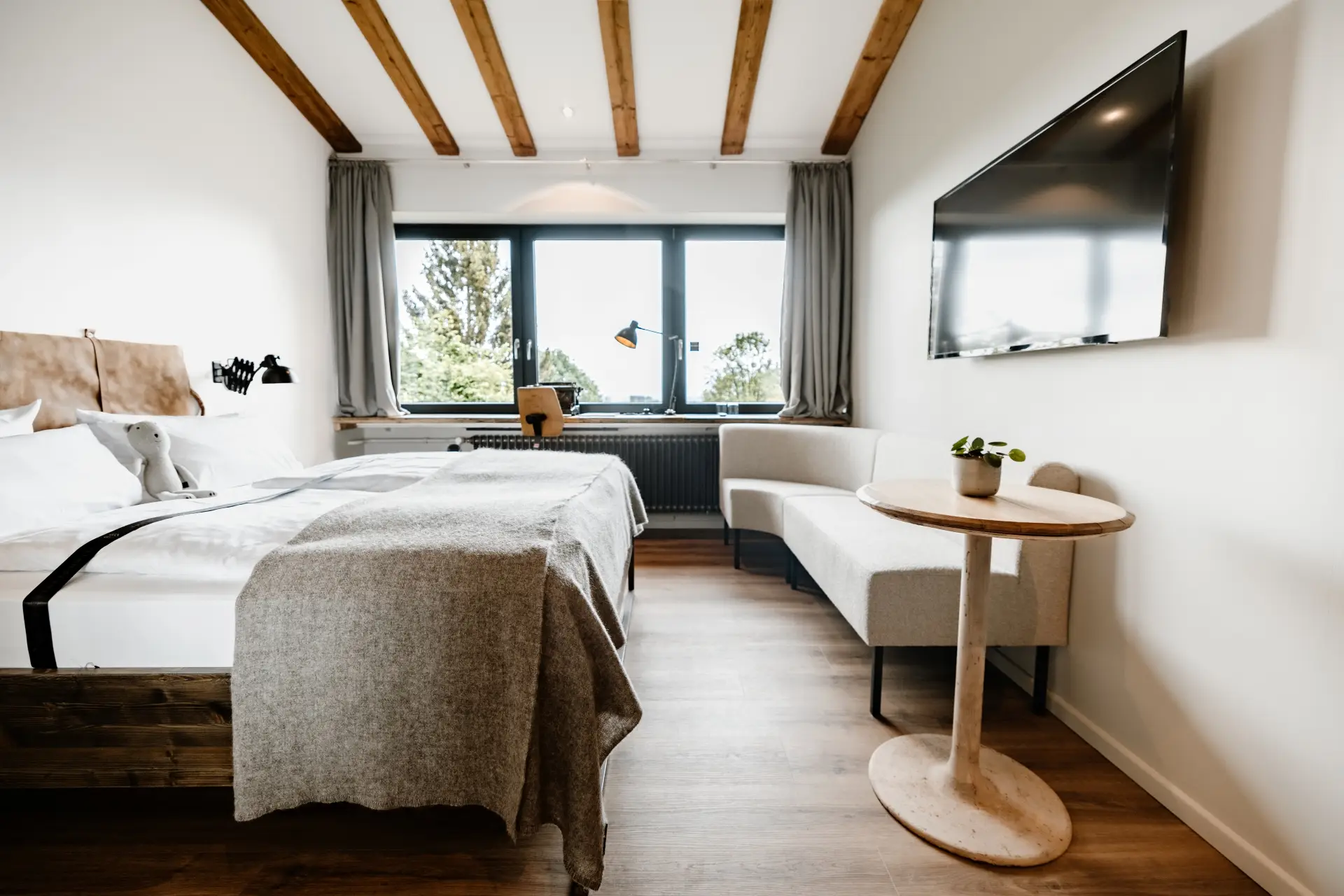 Helles Zimmer mit Doppelbett, Fernseher, Sitzgelegenheit und Fensterblick ins Freie. Plüschtier auf dem Bett.