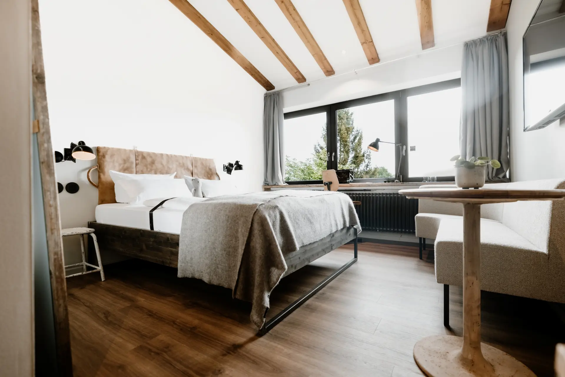 Modernes Schlafzimmer mit einem Doppelbett, Tisch, Stuhl und Fenster zur Außensicht, Holzfußboden und neutraler Farbgebung.