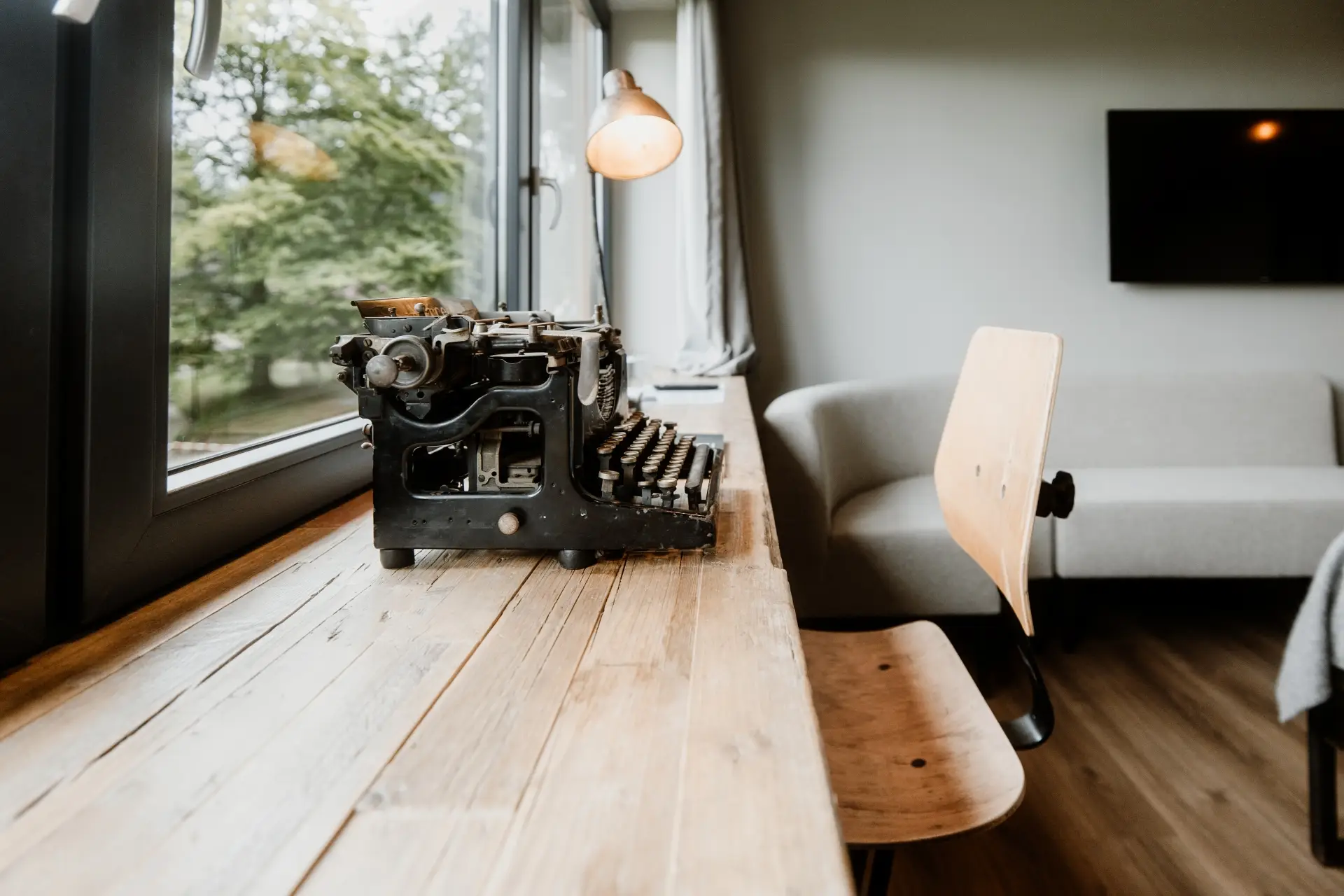 Antike Schreibmaschine auf einem Holztisch, Fenster mit Blick auf den Garten, modernes Interieur mit Sofa und Lampe.
