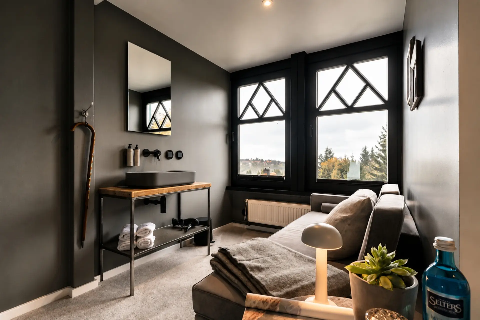 Moderne Ecke eines Zimmers mit grauen Wänden, Waschbecken, Sessel und Blick durch Fenster auf Bäume.