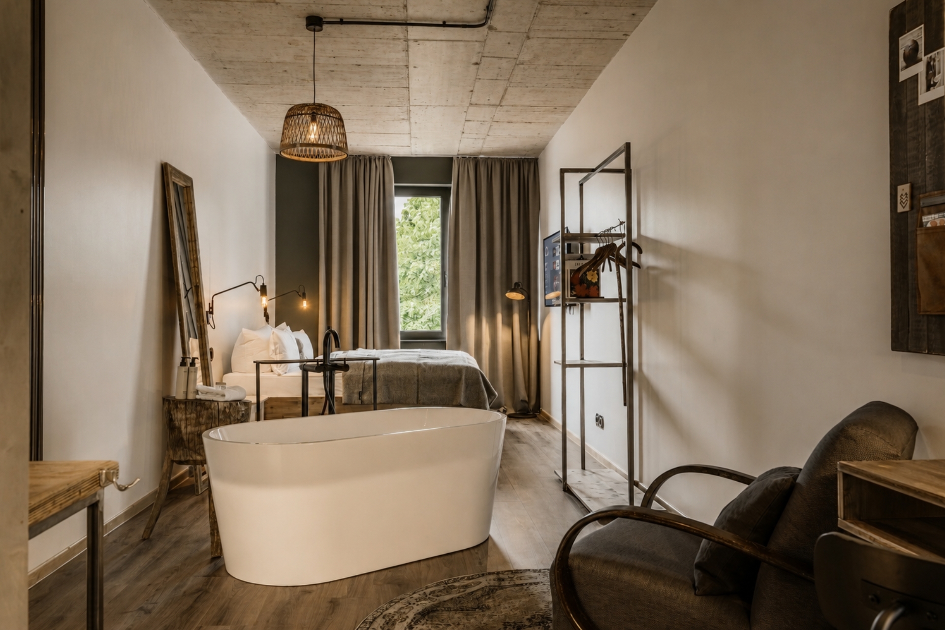 Helles Hotelzimmer mit freistehender Badewanne, Bett, Stuhl und Fenster mit Blick ins Grüne. Moderne Einrichtung mit Holzdetails.
