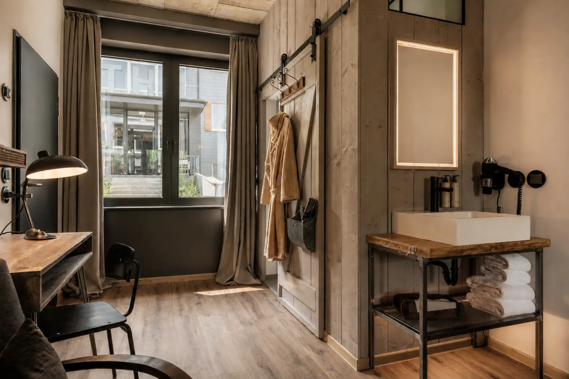 Modernes Zimmer mit Holz- und Betonelementen, Fenstern zur Außenterrasse, Waschbecken und Handtüchern sichtbar.