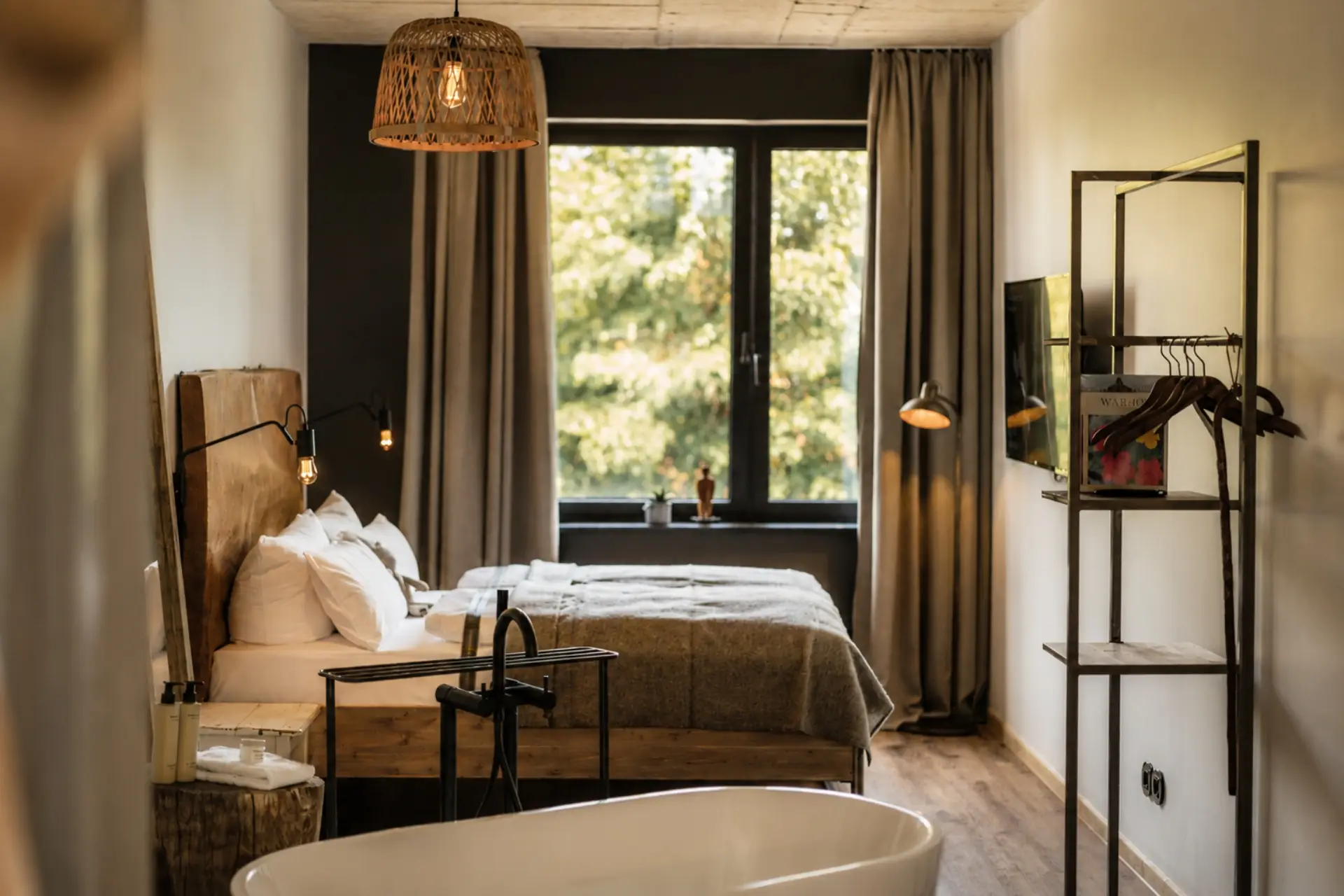 Helles Hotelzimmer mit Doppelbett, freistehender Badewanne und Blick auf einen begrünten Innenhof durch große Fenster.