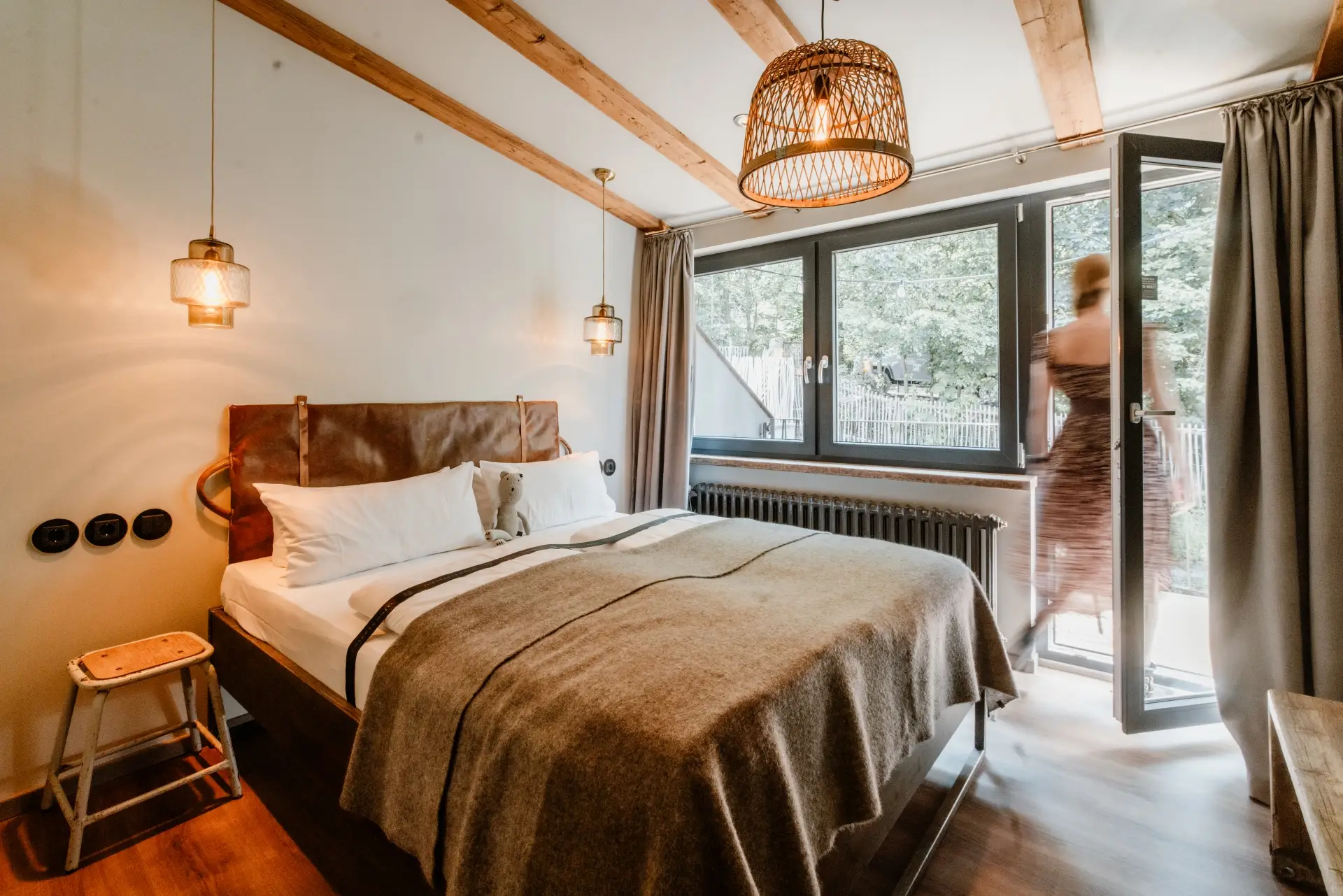 Hellbraunes Schlafzimmer mit einem Bett, Fenster offen, Person verlässt den Raum, Blick ins Grüne. Lichter und dekorative Elemente sichtbar.