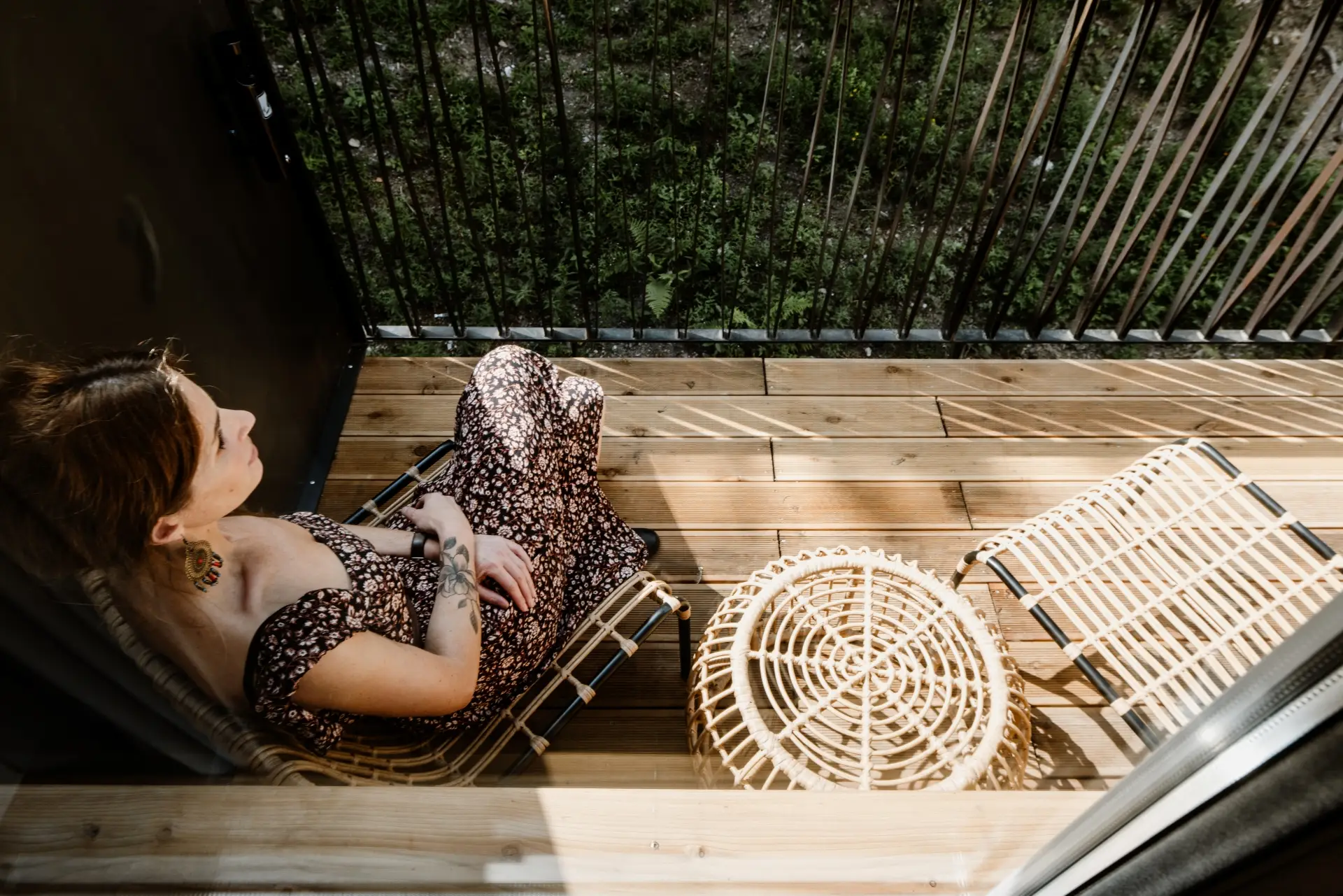 Junge Frau sitzt auf einem Balkon auf Holzlatten, umgeben von zwei Rattanstühlen und Blick ins Grüne.