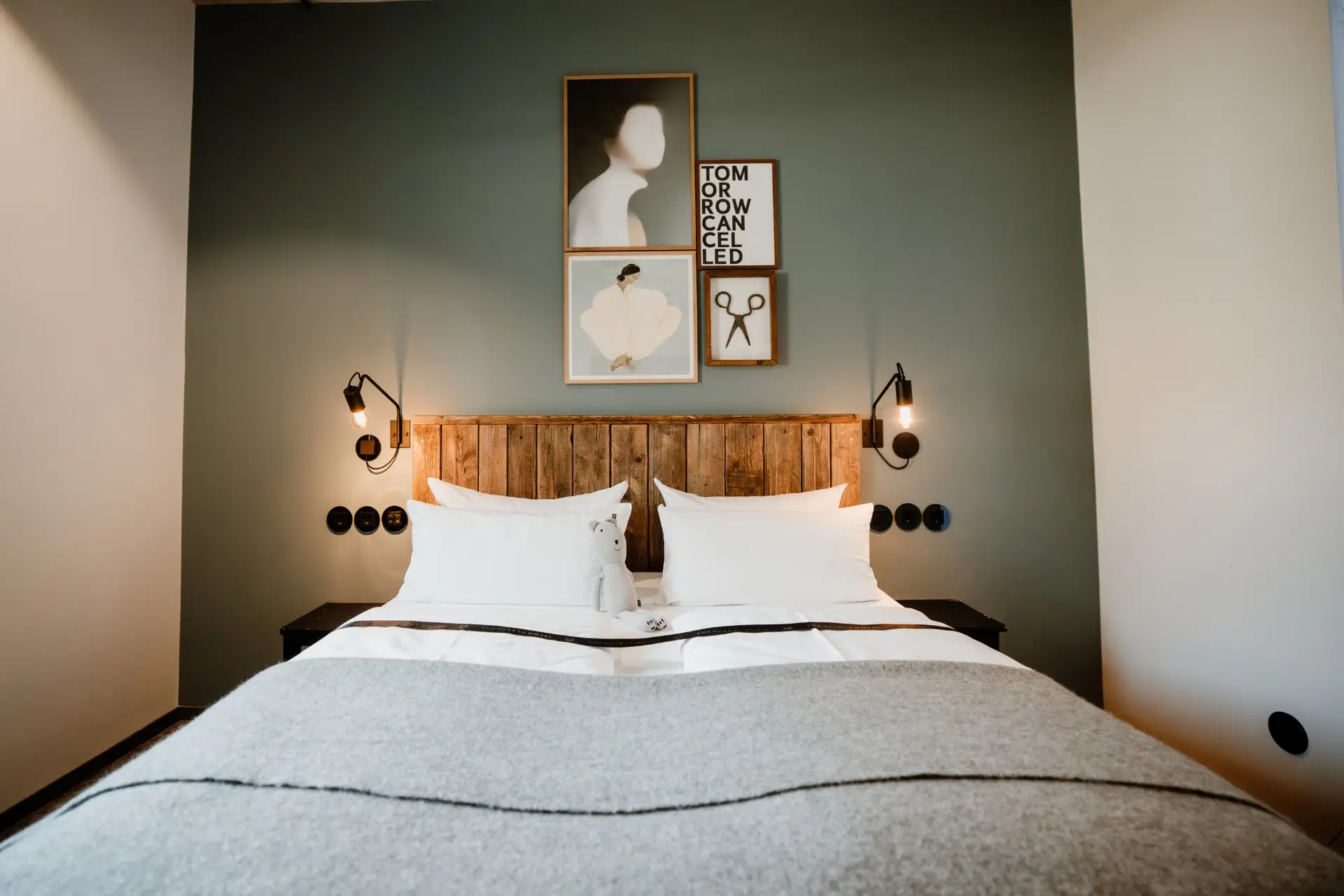 Modernes Schlafzimmer mit Bett, weißen Kissen, Holzkopfleiste, Kunstwerken an der Wand und einem grauen Überwurf.