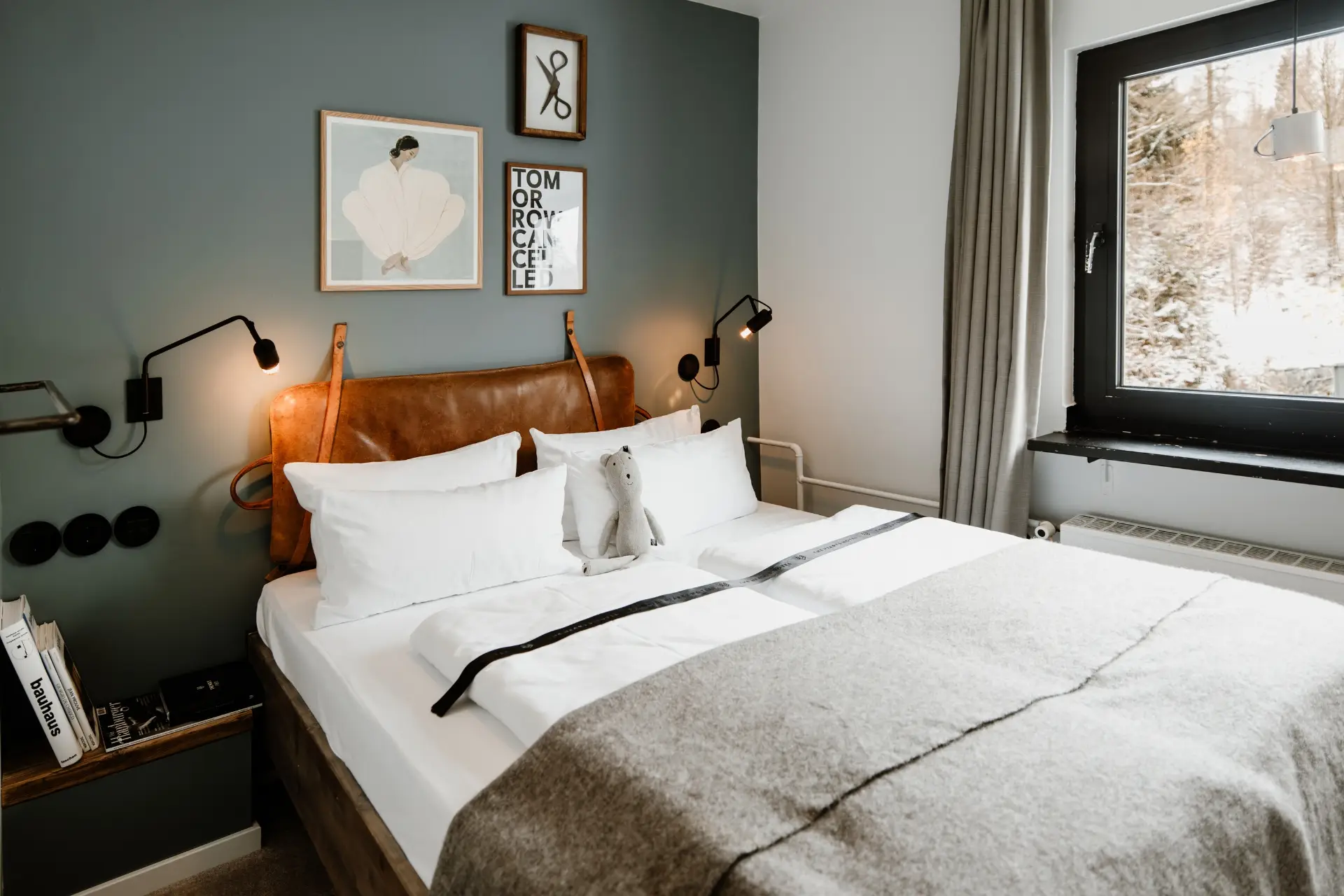 Gemütliches Schlafzimmer mit Doppelbett, weißen Kissen, grauer Decke und dekorativem Plüschtier. Fensterblick auf winterliche Landschaft.