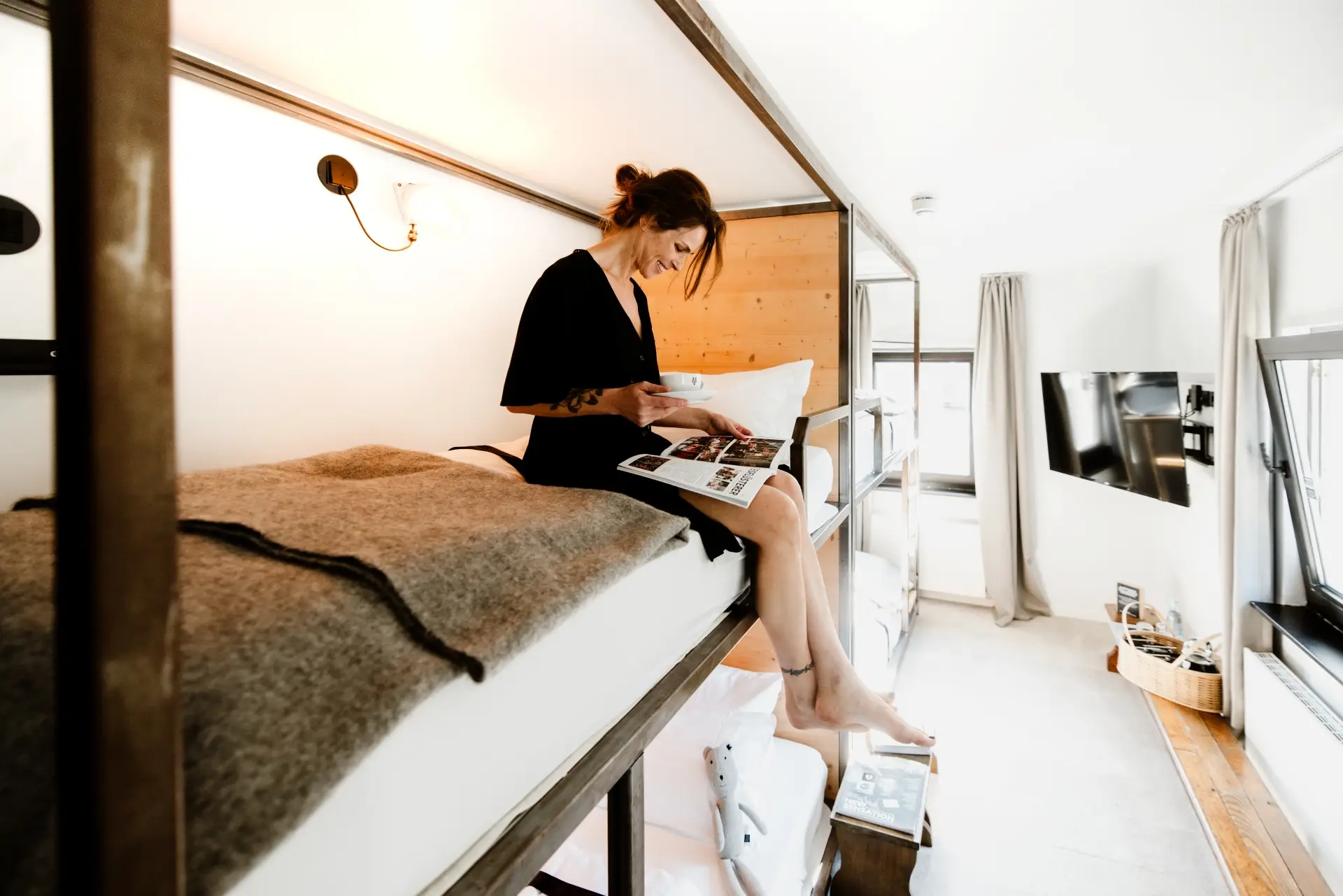 Frau sitzt auf einem Hochbett, liest eine Zeitschrift und genießt einen Kaffee in einem modernen, hellen Zimmer.