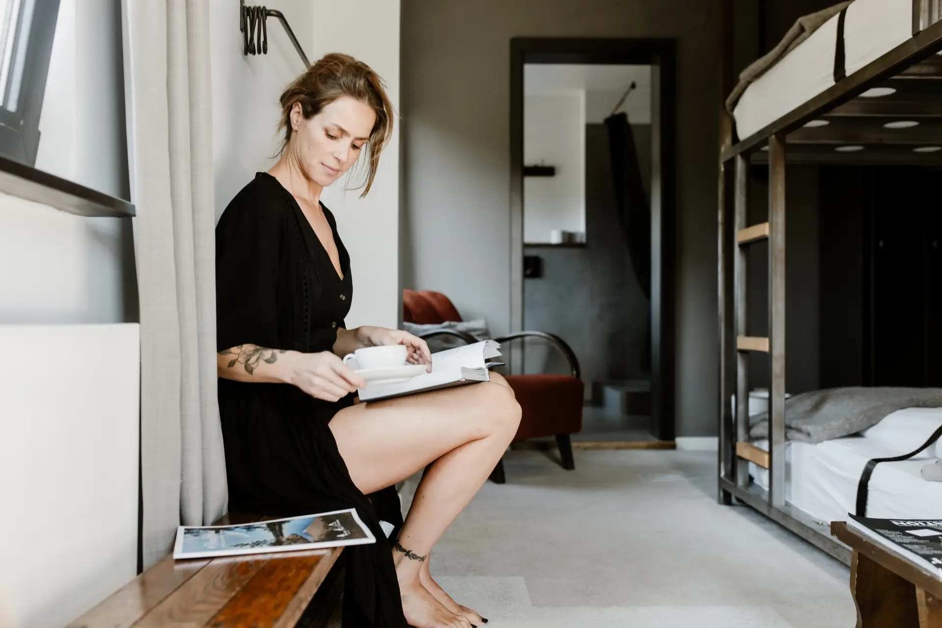Frau sitzt in einem modernen Zimmer, liest ein Buch und hält eine Tasse. Im Hintergrund ist ein Hochbett und eine Sitzgelegenheit sichtbar.