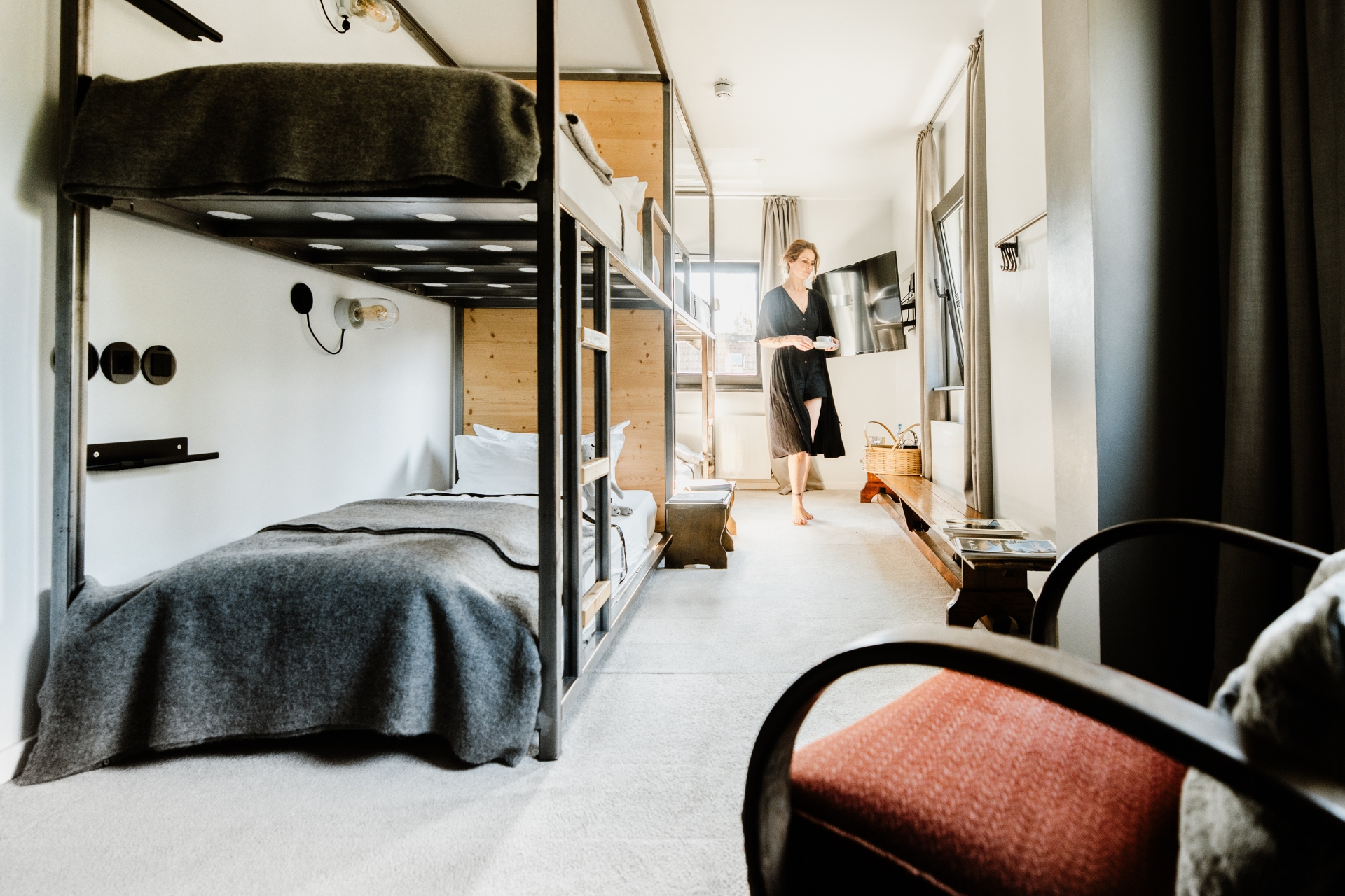 Frau mit Tasse in einem modernen Schlafraum mit Etagenbett, Holzmöbeln und großem Fenster.