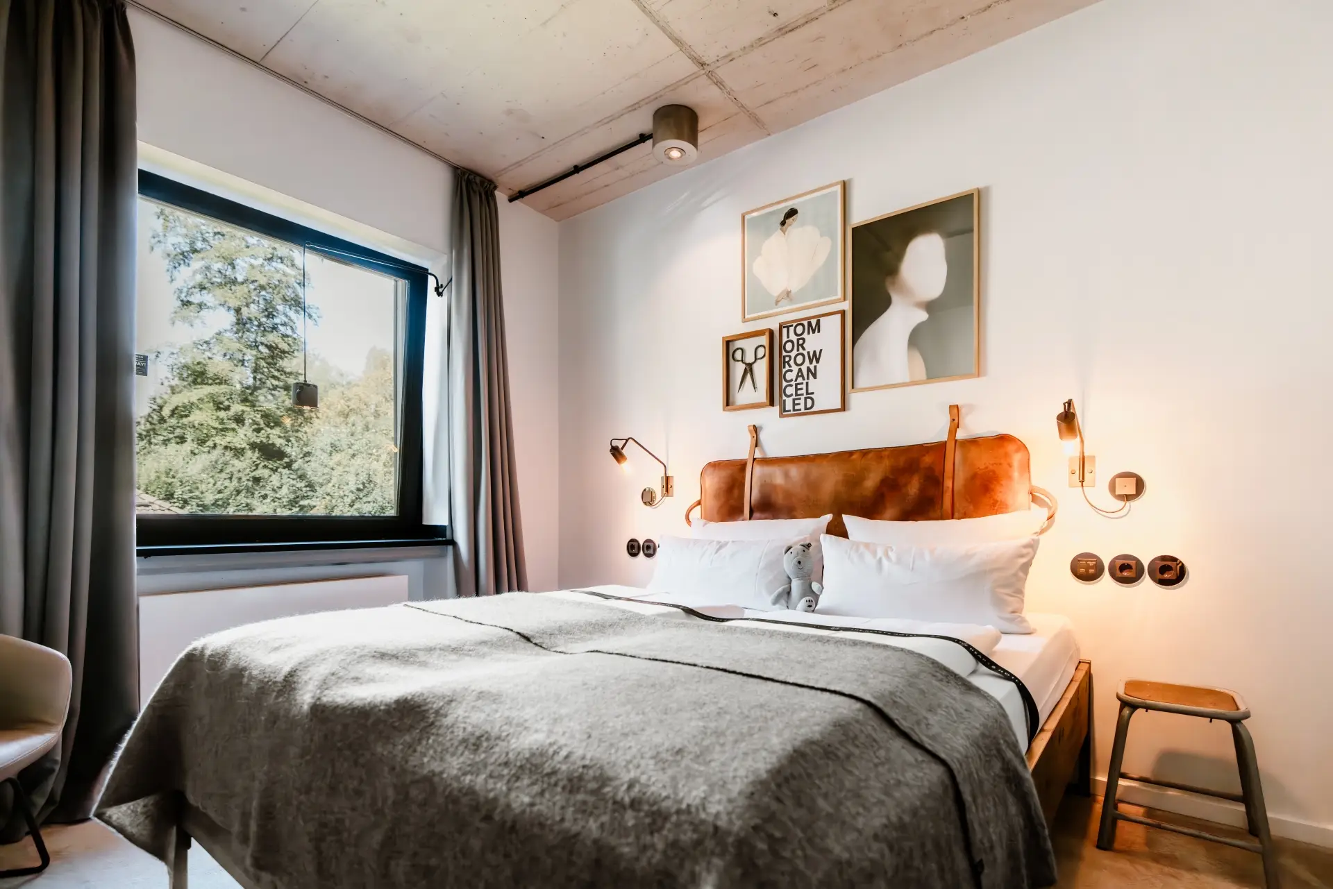 Schlafzimmer mit Doppelbett, grauer Decke und Bildern an der Wand. Fenster mit Blick auf grüne Bäume. Moderne Einrichtung.