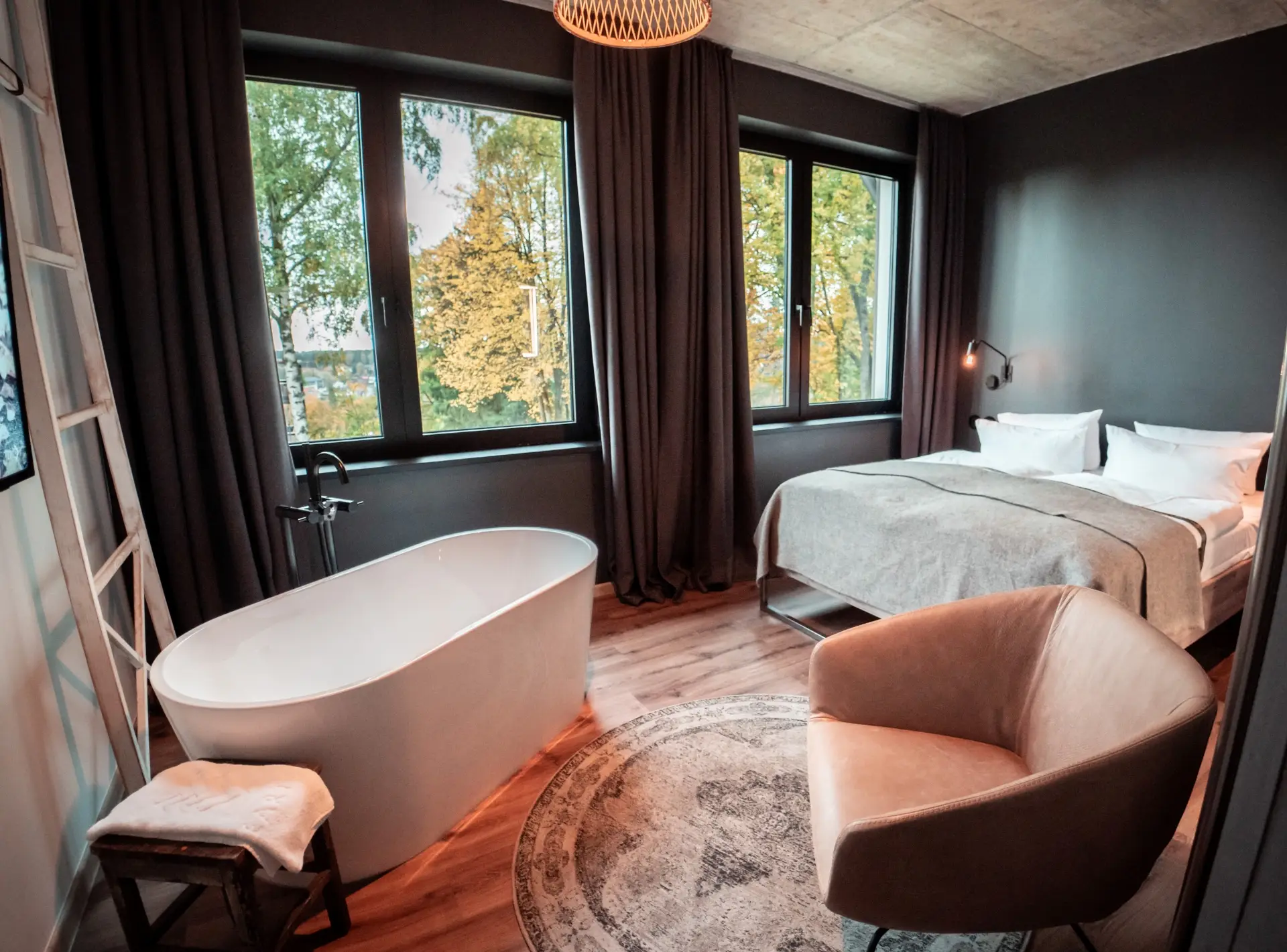 Moderne Hotelzimmer mit Doppelbett, freistehender Badewanne, gemütlichem Sessel und großen Fenstern mit Blick ins Grüne.