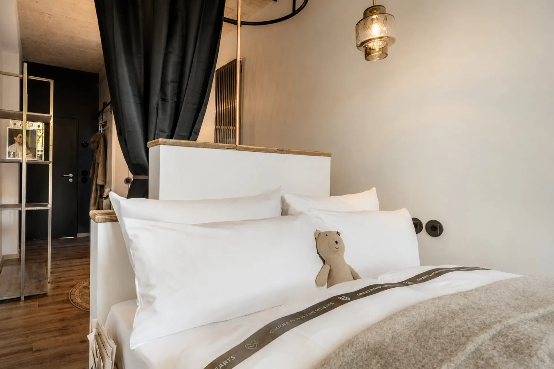 Gemütliches Bett mit weißen Kissen und einem Stoffbär, hinter den Vorhängen ist ein moderner Raum mit Holzdetails.