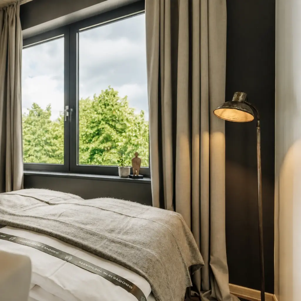 Stimmungsvolles Schlafzimmer mit Fenster, Blick auf Bäume, einem Bett mit grauer Decke und einer Stehlampe.