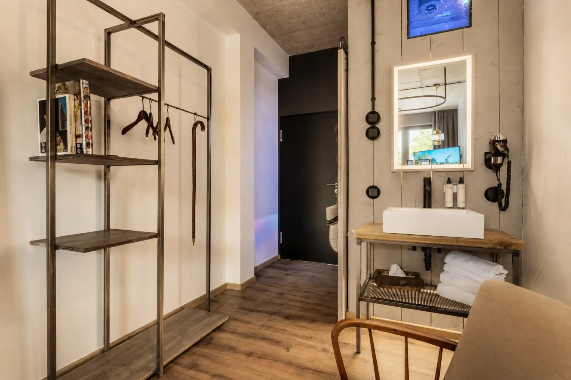 Moderne Hotelzimmer-Ecke mit Regalsystem, Waschbecken, Handtüchern und einem Spiegel. Auf dem Schrank stehen Pflegeprodukte.