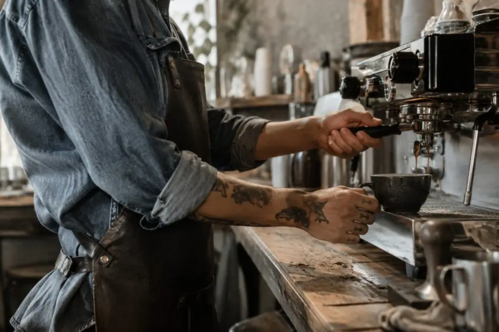 Barista bereitet Kaffee mit einer Espressomaschine an einem Holzarbeitsplatz, tätowierte Hände halten eine Tasse.