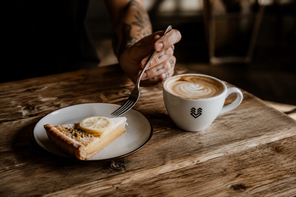 Eine Hand mit Tattoos hält eine Gabel, die in ein Stück Zitronentarte auf einem Teller gestoßen wird, daneben steht eine Tasse Kaffee.