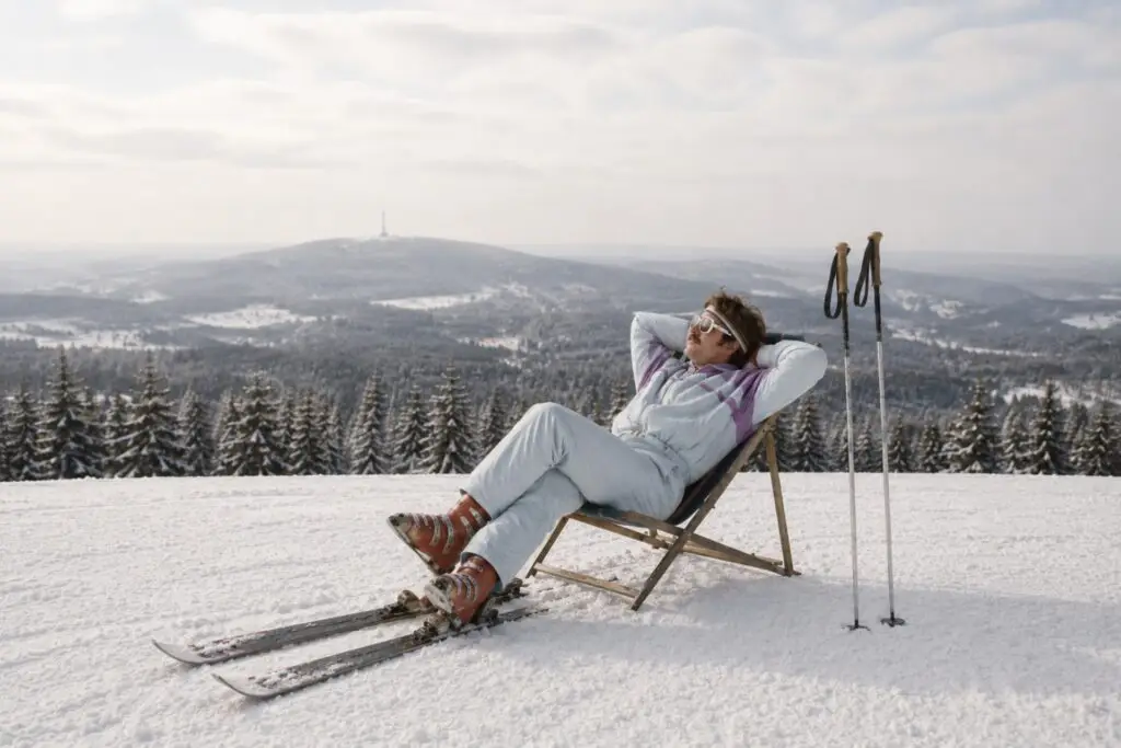 Mann in Skikleidung entspannt auf einem Liegestuhl im Schnee mit verschneiten Bergen und Tannenbäumen im Hintergrund.