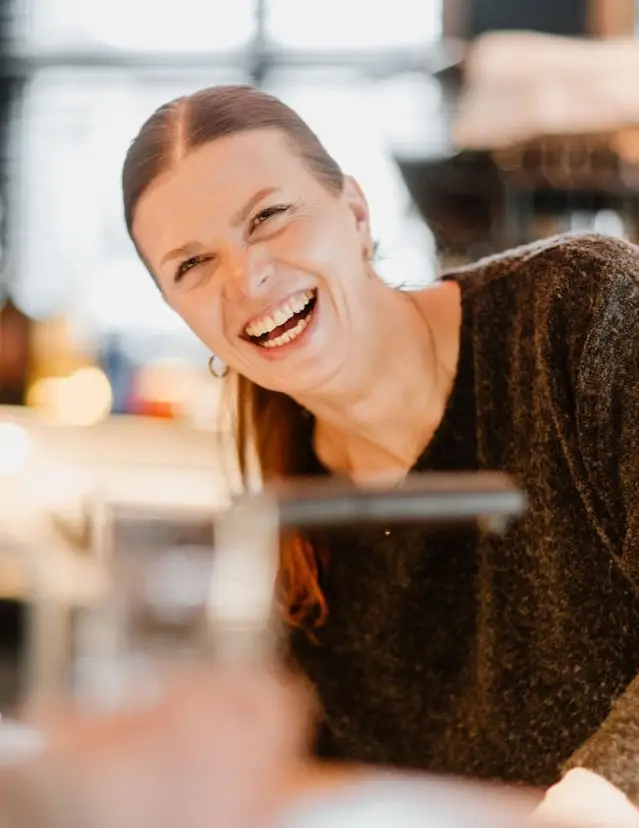 Frau mit langen braunen Haaren lacht herzlich in einem modernen Café, im Hintergrund verschwommene Lichter.
