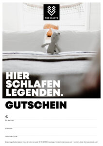 Gutschein für das Hotel The Hearts in Braunlage, mit einem Stofftier auf einem Bett zwischen Kissen.