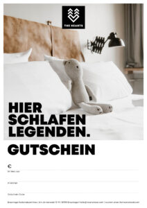 Alt-Text: Gutschein für das Hotel The Hearts liegt auf einem Bett mit Kissen und einem Stofftier in Form eines Bären.