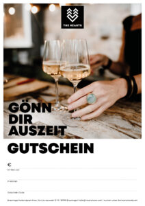 Zwei Gläser Sekt stehen auf einem Tisch, während eine Hand mit einem Ring einen Gutschein für Auszeit hält.
