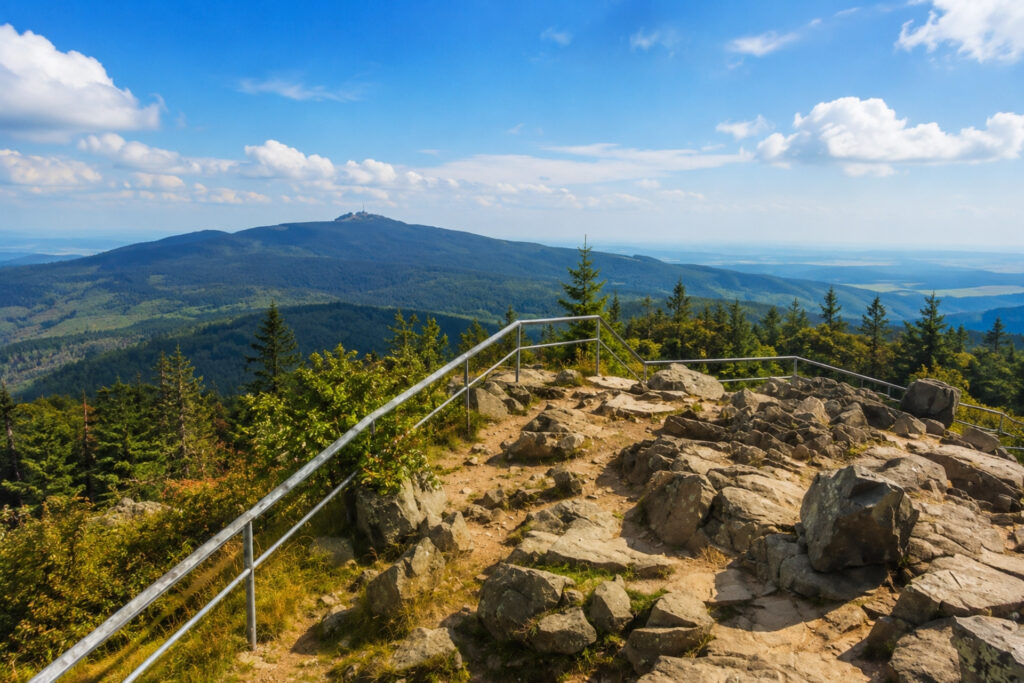 Aussichtspunkt mit Geländer und Felsen. Im Hintergrund erstrecken sich Wälder und Berge unter einem blauen Himmel mit wenigen Wolken.
