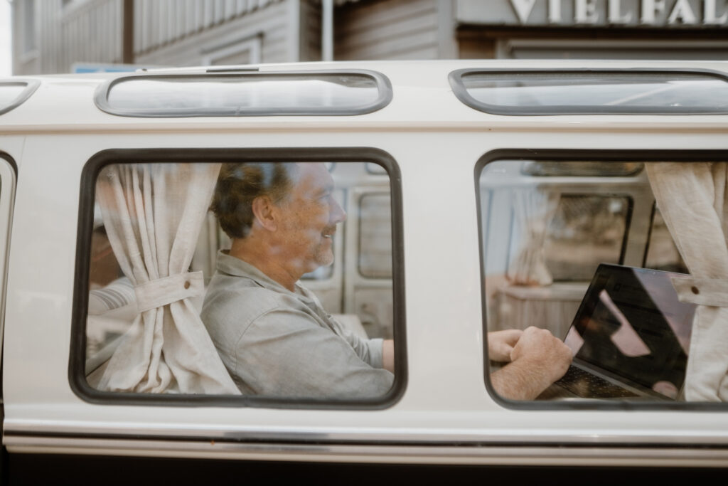 Mann sitzt in einem VW-Bus und arbeitet an einem Laptop, während er entspannt durch das Fenster lächelt.