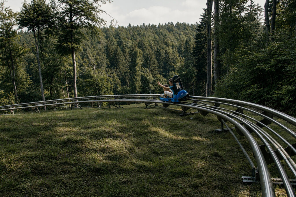 Ein Roller mit einer Person fährt auf einer kurvigen Gleisbahn durch eine grüne Landschaft mit Bäumen im Hintergrund.