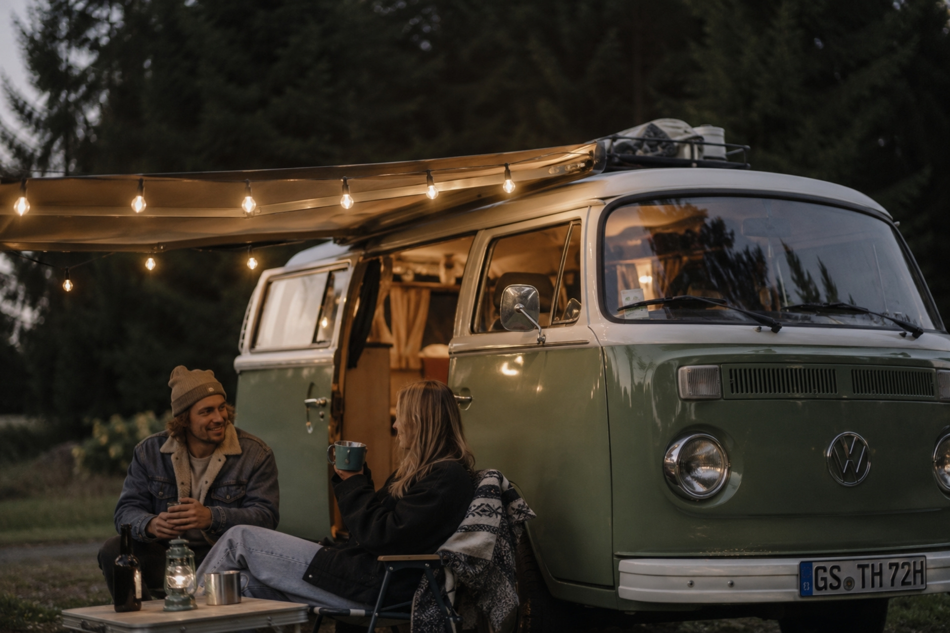 Paar sitzt vor einem VW-Bus im Freien, trinkt aus Tassen und genießt die Abendstimmung bei lampenbeleuchtetem Campingplatz.