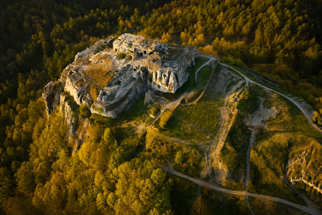 Luftaufnahme einer alten Felsenburg, umgeben von dichten Wäldern, mit visiblem Wegenetz und grünen Flächen.