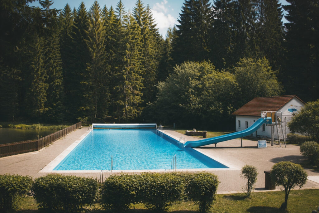 Blauer Swimmingpool mit Wasserrutsche, umgeben von Bäumen, und einem kleinen Gebäude im Hintergrund. Klarer blauer Himmel.
