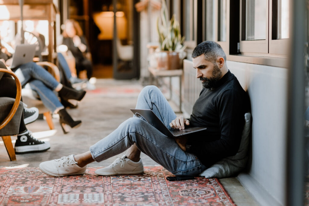 Mann sitzt auf dem Boden, arbeitet an Laptop. Im Hintergrund sind andere Personen in einem modernen Innenraum.
