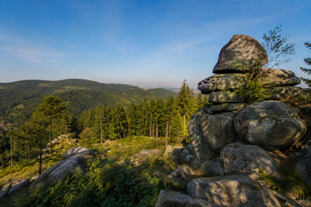 Felsformation mit mehreren großen Steinen, umgeben von Wald und sanften Hügeln, unter einem klaren blauen Himmel.