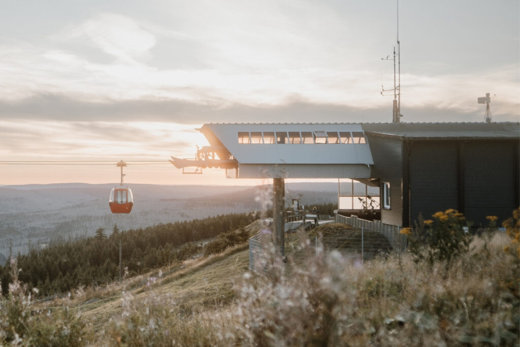 Seilbahnstation auf einem Hügel, mit roter Gondel und Sonnenuntergang über bewaldeter Landschaft im Hintergrund.
